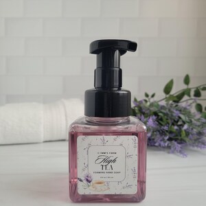 May include: A clear, square bottle of High Tea foaming hand soap with a black pump dispenser. The pink liquid is visible through the bottle. The label features the text "High Tea" and a teacup illustration. The background includes a white towel and lavender sprigs.