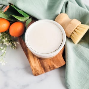 May include: A round white ceramic dish filled with a solid white cleaning paste, accompanied by a wooden scrub brush with light-colored bristles. The set is arranged on a small wooden board next to two small orange fruits and green leaves.