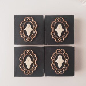 May include: Four black square soaps with a gold-colored ornate frame design. Each frame contains a white ghost figure. The soaps are arranged in a 2x2 grid, perfect for Halloween or spooky-themed decor.