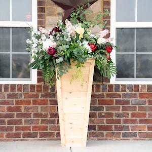 Puede incluir: Un macetero alto de madera cónico lleno de una variedad de flores en tonos rojos, rosas y blancos. El macetero está frente a una pared de ladrillo con dos ventanas.