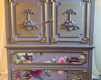 May include: A vintage-style cabinet with a lavender paint finish and gold accents. The cabinet has two doors with ornate details and two drawers with floral patterned paper.