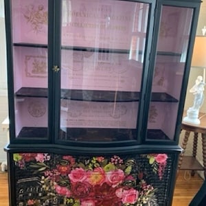 May include: A black china cabinet with glass doors and shelves. The cabinet has a floral design on the bottom drawers. The glass doors have a pink background with gold lettering that reads "Botanical Bazaar Collection of Herbs".