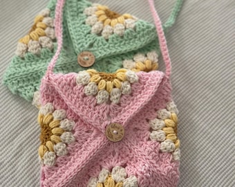 Pode incluir: Duas bolsas de crochê com desenhos florais. A bolsa menor é rosa com flores amarelas e brancas e um botão de madeira que diz "Woven & Knit". A bolsa maior é verde com flores brancas e amarelas.