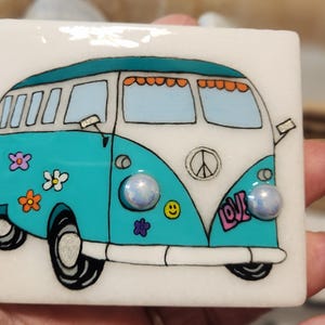 May include: A square, white ceramic tile featuring a colorful illustration of a teal and white vintage van. The van has a peace sign, the word "LOVE", and flower and smiley face details. Two iridescent, round embellishments are on the van.