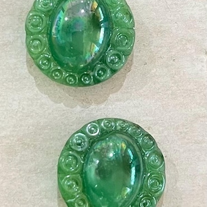 May include: Two green, oval buttons with a swirl pattern around the edges. The buttons have a glossy, iridescent finish, reflecting light. These buttons are suitable for sewing or craft projects.