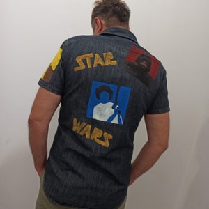 以下が含まれることがあります： 後ろに金色の「スターウォーズ」のデザインが施された濃い青色のデニムシャツ。デザインには、レイア姫とダースベイダーのシルエットが含まれています。