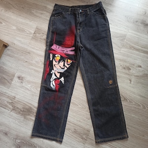 以下が含まれることがあります： 赤い帽子、白いシャツ、赤いネクタイを着た漫画のキャラクターが手描きで描かれた黒いデニムのジーンズ。キャラクターは不気味な笑顔で、タバコをくわえている。