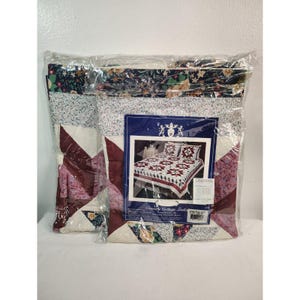 May include: A packaged Country Cottage Quilt set. The quilt features a patchwork design with burgundy, cream, and floral patterns. The packaging includes a picture of the quilt on a bed with matching pillow shams.