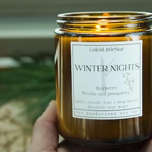 Puede incluir: Una vela en tarro de cristal marrón con una etiqueta blanca que dice "WINTER NIGHTS" y "Bayberry". La vela es de cera de soja vertida a mano y tiene una tapa dorada. La vela se sostiene en una mano.