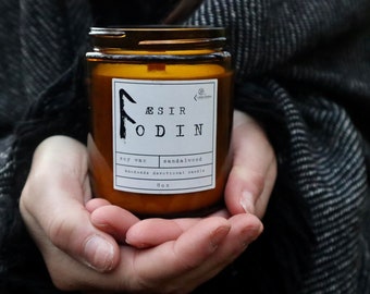 May include: A brown glass jar candle with a black lid, held in cupped hands. The label reads "ÆSIR ODIN" with "soy wax" and "sandalwood" details. The candle is 8 oz.