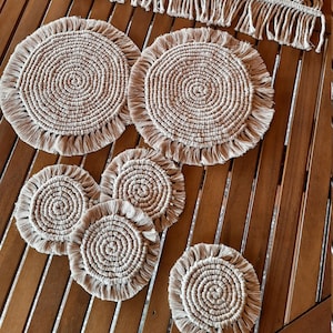 May include: Set of six beige crocheted coasters with fringe. The coasters are arranged on a wooden table with a beige crocheted table runner.