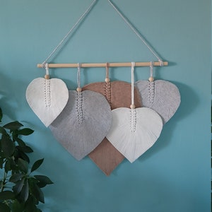 Puede incluir: Un tapiz de pared con cuatro piezas de macramé en forma de corazón en tonos grises, blancos y marrones. Los corazones están dispuestos en forma escalonada, con el corazón más grande en la parte inferior y el más pequeño en la parte superior. Los corazones están hechos con un hilo grueso y texturizado y tienen una franja en la parte inferior.