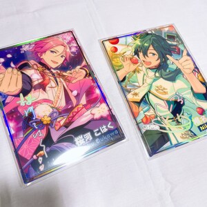 May include: Two holographic cards featuring anime characters. The card on the left features a pink-haired character with a pink and white outfit. The card on the right features a green-haired character with a white and green outfit. The text on the left card reads "桜河 こはく"