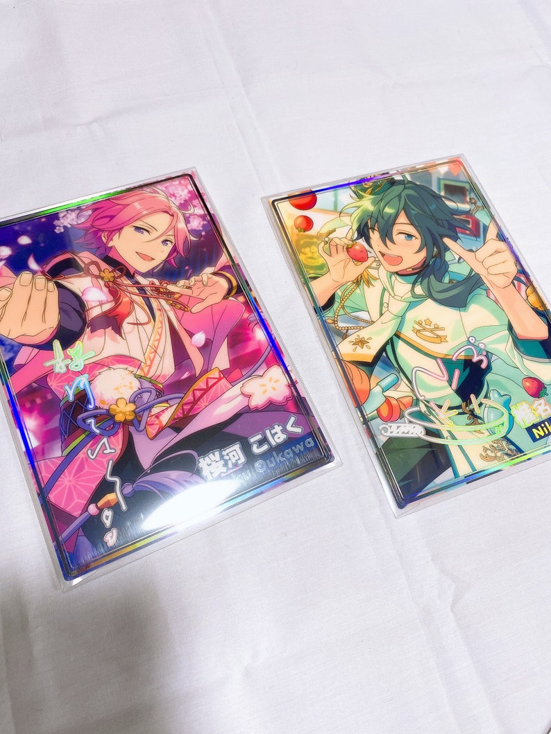 May include: Two holographic cards featuring anime characters. The card on the left features a pink-haired character with a pink and white outfit. The card on the right features a green-haired character with a white and green outfit. The text on the left card reads "桜河 こはく"