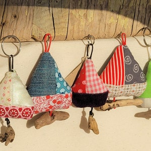 Könnte beinhalten: Fünf bunte Stoff-Segelboot-Schlüsselanhänger mit Treibholz-Akzenten. Jeder Schlüsselanhänger hat ein anderes Patchwork-Design mit roten, weißen, blauen, grünen und grauen Stoffen.