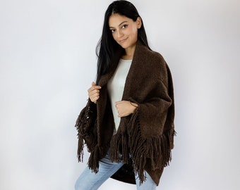 May include: A brown knitted shawl with fringe trim. The shawl is worn over a white shirt and blue jeans. The model is wearing brown leather boots.