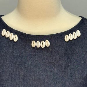 May include: Dark blue denim top with a scoop neckline. The neckline is adorned with small, white seashells arranged in clusters. The top is displayed on a mannequin.