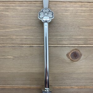May include: A silver metal straw with a decorative flower design at the top and a spiral design at the bottom.