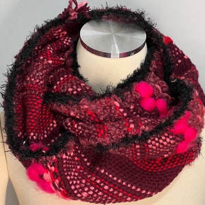 Puede incluir: Una bufanda infinita de color burdeos y rosa con flecos negros y pompones rosas.