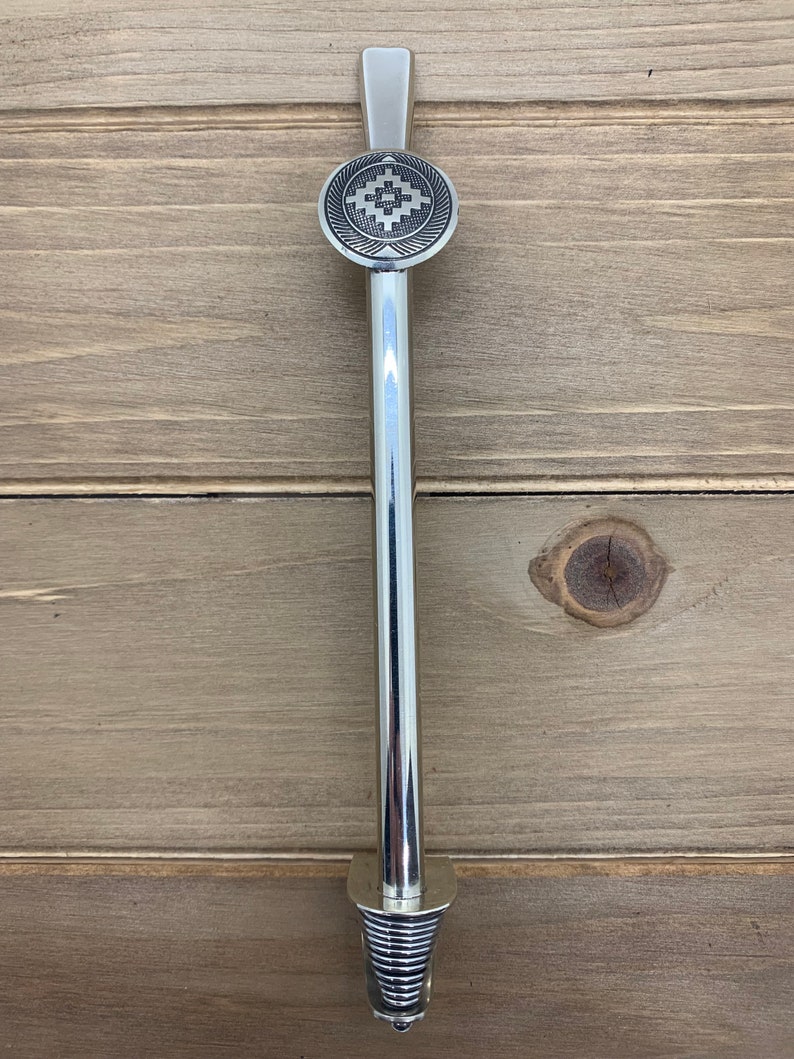 May include: A silver metal stirring stick with a decorative round top featuring a geometric design. The stick has a spiral base.