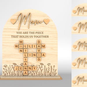 以下が含まれることがあります： 「ママ」という言葉と「あなたは私たちをまとめるピースです」というテキストが書かれた木製の看板。看板には、スクラブルタイルのクロスワードパズルがあり、「ウィリアム」、「リニア」、「ジェニー」という名前が書かれています。