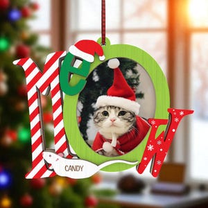 May include: A Christmas ornament featuring a cat wearing a Santa hat and red coat. The ornament spells out "MEOW" with red and white striped letters and green letters. The name "Candy" is written on a white mouse.