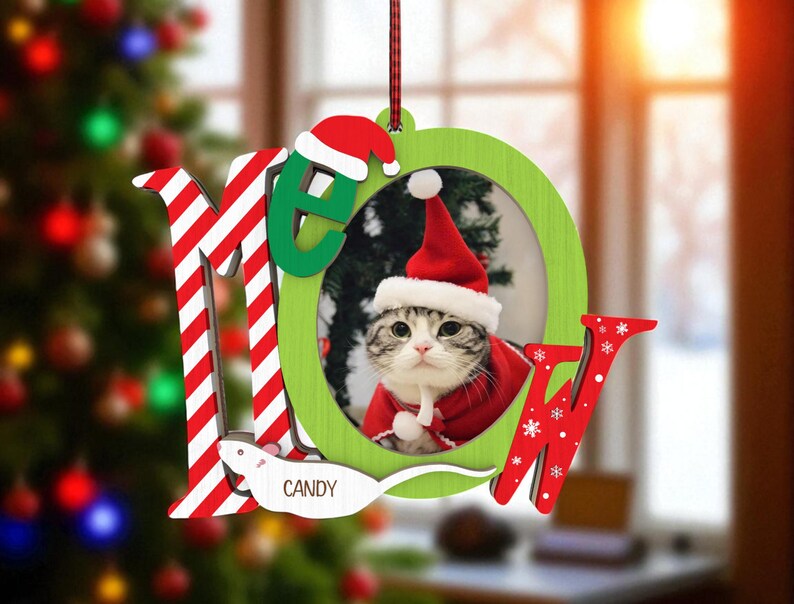 May include: A Christmas ornament featuring a cat wearing a Santa hat and red coat. The ornament spells out "MEOW" with red and white striped letters and green letters. The name "Candy" is written on a white mouse.