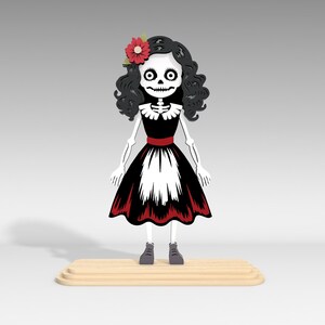 Pu&ograve; includere: Una figura stilizzata di scheletro in un vestito nero e bianco con rifiniture rosse e un fiore rosso tra i capelli. La figura ha capelli neri ricci, un volto scheletrico e si trova su una piattaforma di legno. La figura indossa scarpe grigie.