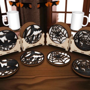 Peut inclure: Ensemble de dessous de verre sur le thème d'Halloween avec des motifs découpés au laser de chauves-souris, de toiles d'araignées et d'une maison hantée. Les dessous de verre sont bruns avec des accents blancs et noirs, présentés dans des supports en bois. Deux mugs blancs sont également visibles.