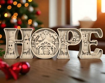 May include: Wooden "HOPE" sign with a nativity scene in the "O". The letters are light-colored and feature cut-out designs of buildings, animals, and stars. A Christmas tree with red and gold ornaments is in the background.