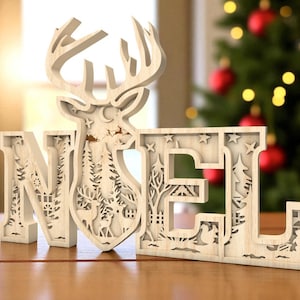 May include: Wooden Christmas decoration spelling "NOEL" with a reindeer head and a winter scene cut out within the letters. The letters are light-colored and sit on a wooden surface, with a Christmas tree in the background.