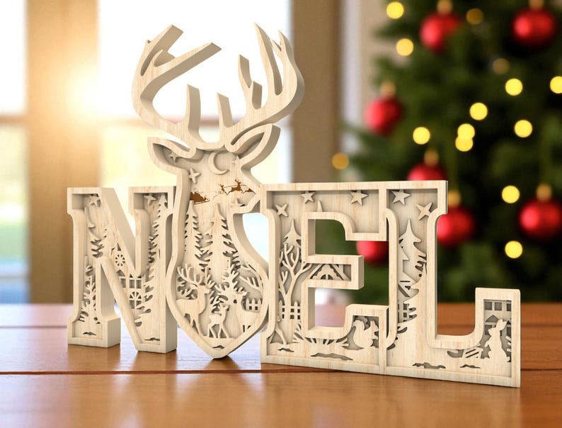 May include: Wooden Christmas decoration spelling "NOEL" with a reindeer head and a winter scene cut out within the letters. The letters are light-colored and sit on a wooden surface, with a Christmas tree in the background.