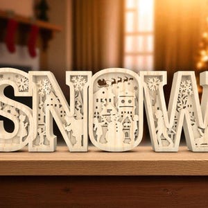 May include: Wooden "SNOW" sign with detailed cut-out designs. The letters feature winter scenes, including snowflakes, houses, and figures. The sign is made of light-coloured wood and sits on a wooden surface.