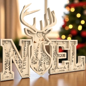 May include: A wooden "NOEL" Christmas decoration with a deer silhouette and forest scene cutouts. The letters are in a light wood color, with intricate details of trees, stars, and a house. The background is blurred, with a Christmas tree.