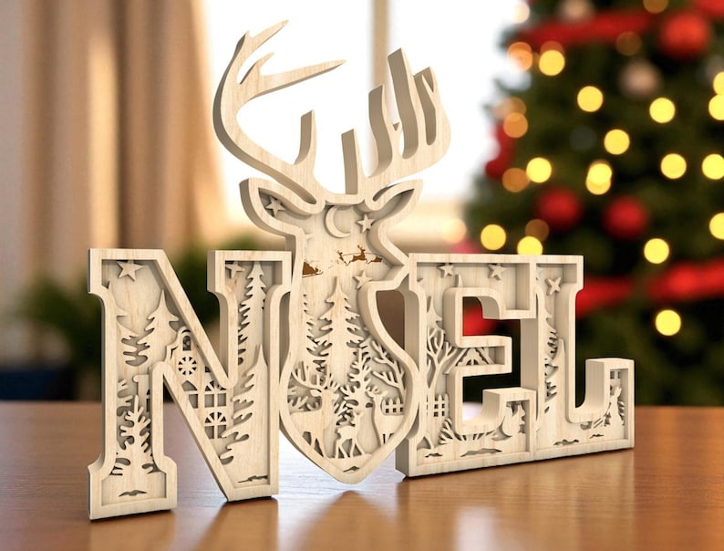 May include: A wooden "NOEL" Christmas decoration with a deer silhouette and forest scene cutouts. The letters are in a light wood color, with intricate details of trees, stars, and a house. The background is blurred, with a Christmas tree.