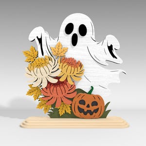 Pu&ograve; includere: Decorazione di Halloween in legno con un fantasma bianco con occhi e bocca neri, accompagnato da fiori arancioni e gialli, una zucca intagliata e foglie verdi. La decorazione &egrave; posta su una base di legno.