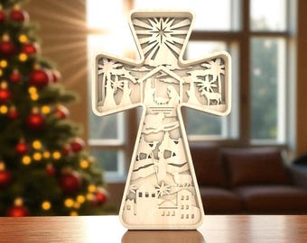 May include: Wooden cross-shaped Christmas decoration with a layered design. The cross features a nativity scene with palm trees, figures, and a starburst at the top. The cross is made of light-colored wood and is displayed on a wooden surface.