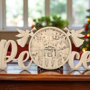 May include: Wooden Christmas decoration with the word "Peace" in cursive script. The letter "e" is connected to a circular cutout depicting a nativity scene, flanked by angel figures. The piece is made of light-colored wood and sits on a wooden surface.