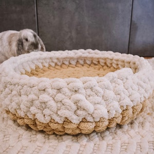 May include: A white and beige knitted pet bed with a fluffy texture. The bed is round and has a raised edge.