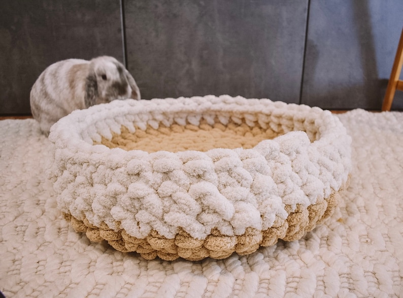 May include: A white and beige knitted pet bed with a fluffy texture. The bed is round and has a raised edge.