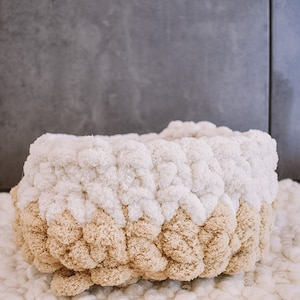 May include: A white and beige knitted basket with a pom-pom texture. The basket is made of yarn and has a round shape.