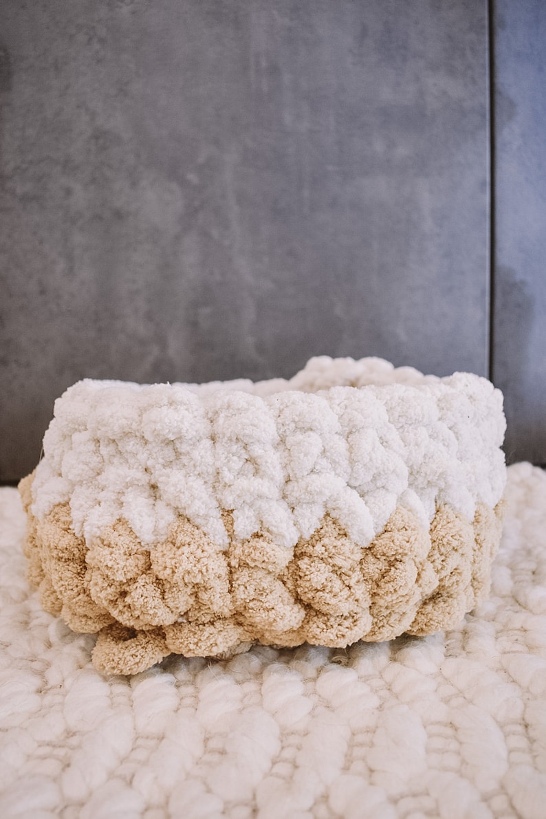May include: A white and beige knitted basket with a pom-pom texture. The basket is made of yarn and has a round shape.