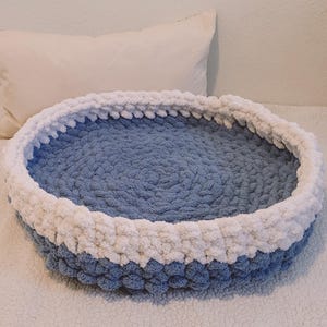 May include: A blue and white crocheted pet bed. The bed is round and has a white rim.