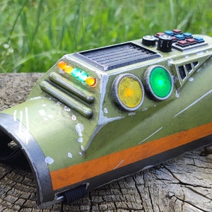 Op de afbeelding: Een futuristisch apparaat in groen en oranje met een reeks lampjes en knoppen. Het apparaat heeft een versleten, verweerde uitstraling, wat suggereert dat het in een ruwe omgeving is gebruikt.