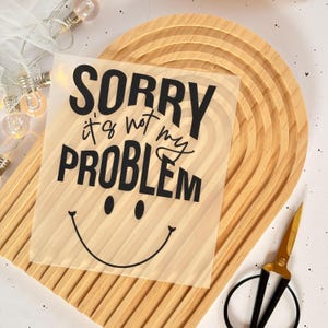 Könnte beinhalten: Ein transparenter Vinyl-Aufkleber mit schwarzem Text, der "Sorry, ist nicht mein Problem" lautet, mit einem Smiley darunter. Der Aufkleber befindet sich auf einer Holzoberfläche.
