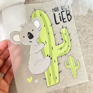Könnte beinhalten: Ein grauer Koalabär umarmt einen grünen Kaktus mit dem Text "Hab Dich Lieb" auf weißem Hintergrund.