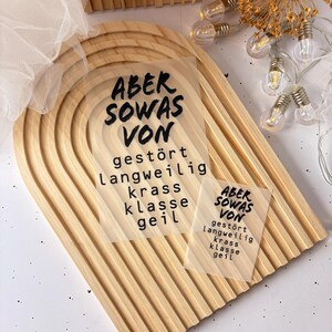 Könnte beinhalten: Zwei transparente Aufkleber mit schwarzem Text auf einer Holzoberfläche. Der Text auf den Aufklebern lautet "ABER SOWAS VON" und enthält die Wörter "gestört", "langweilig", "krass", "klasse" und "geil".