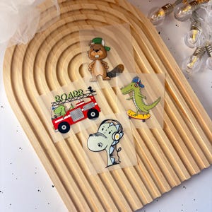 Könnte beinhalten: Vier Bügelbilder mit Comic-Tieren und einem Feuerwehrauto. Die Patches zeigen einen Biber mit grünem Hut, einen Dinosaurier auf einem Skateboard, einen Dinosaurier mit Kopfhörern und ein rotes Feuerwehrauto mit einem Dinosaurier an der Seite. Das Feuerwehrauto hat den Schriftzug "Roar" an der Seite.