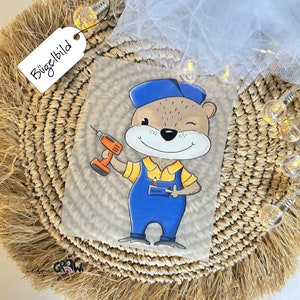 Könnte beinhalten: Bügelbild mit einer Cartoon-Illustration eines braunen Bären, der eine blaue Mütze und eine Latzhose trägt und eine Bohrmaschine hält. Der Bär zwinkert und lächelt. Das Bügelbild ist auf einem transparenten Hintergrund.