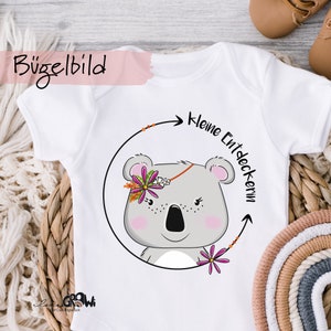 Könnte beinhalten: Weißer Baby Strampler mit einem grauen Koala, der eine rosa Blumenkrone trägt. Der Strampler hat den Text "Kleine Entdeckerin" in einem schwarzen Kreis mit einem Pfeil.